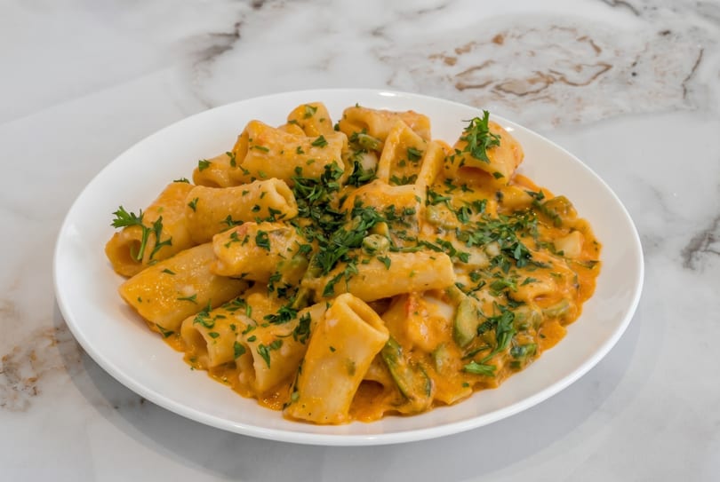 Rigatoni pasta in a creamy orange sauce with green vegetables and fresh parsley on a white plate.