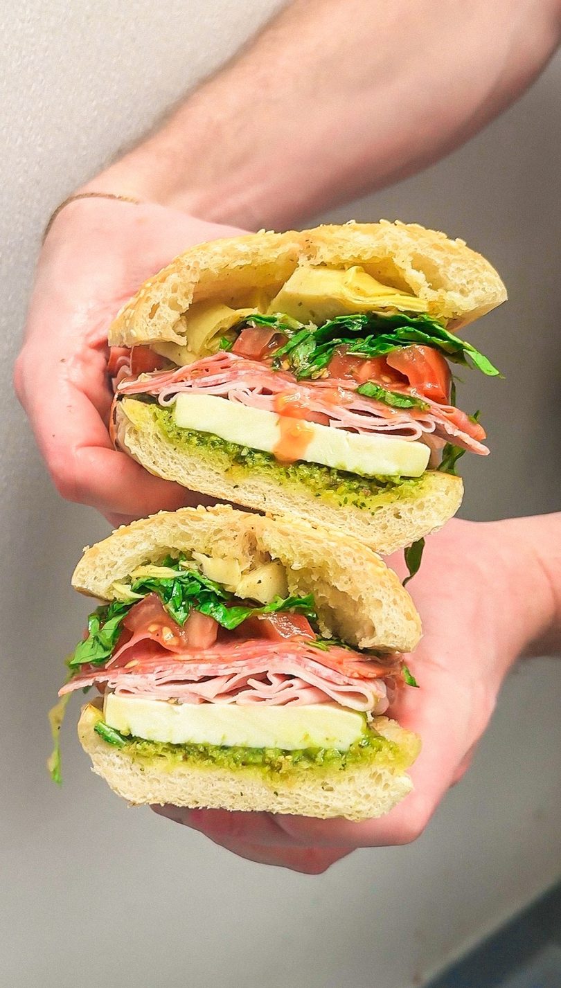 Hands holding a large Italian sandwich, cut in half, filled with meats, cheese, and vegetables.