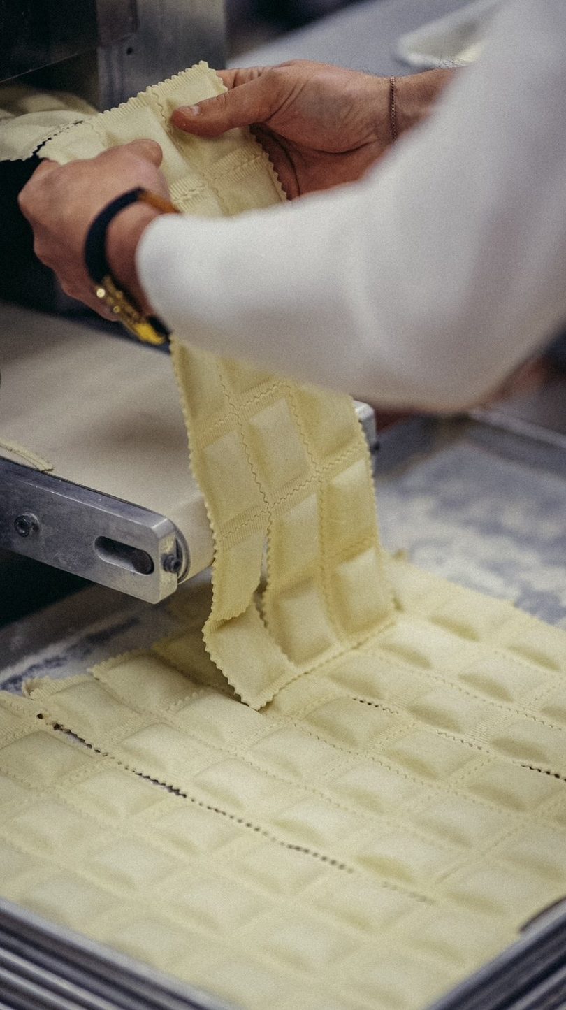 Hands carefully separate freshly made ravioli from a pasta machine, showing rows of square pasta.