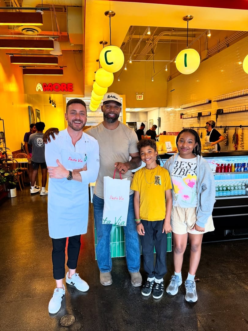 A chef, a man, and two children pose with a Pasta Via bag in a brightly lit restaurant.