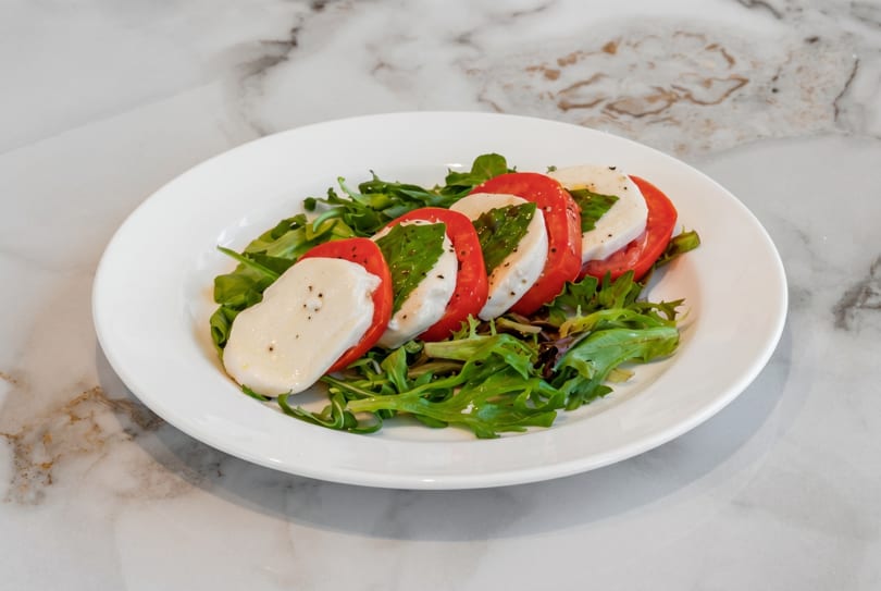 Caprese salad with sliced tomatoes, mozzarella, basil, and arugula on a white plate.