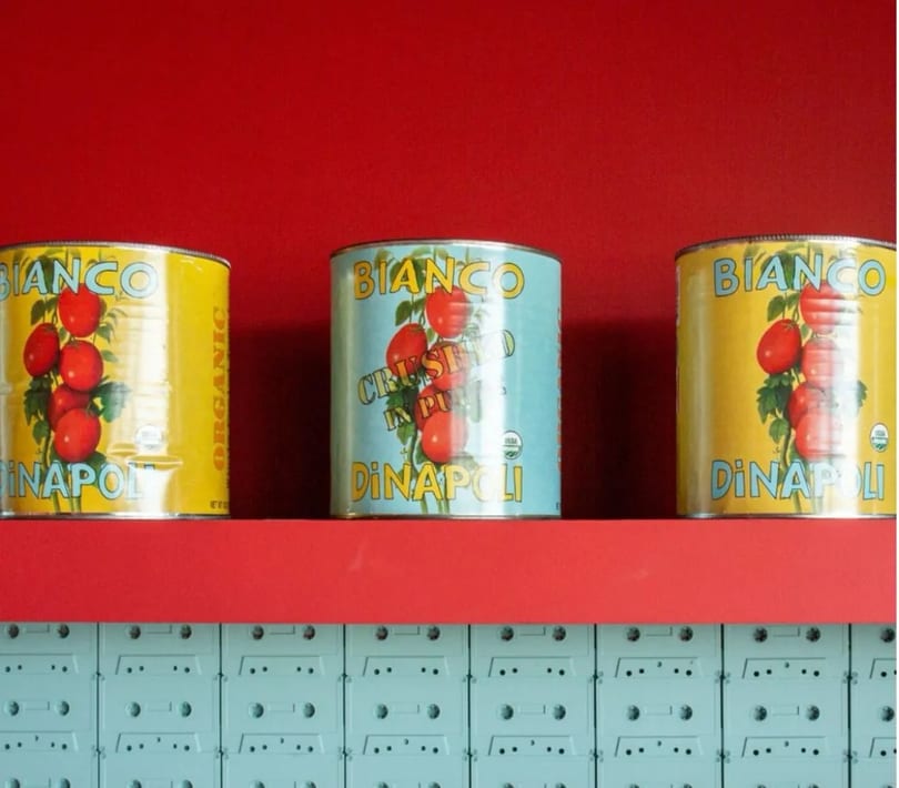 Three colorful Bianco DiNapoli tomato cans on a red shelf above a wall of light blue cassette tapes.