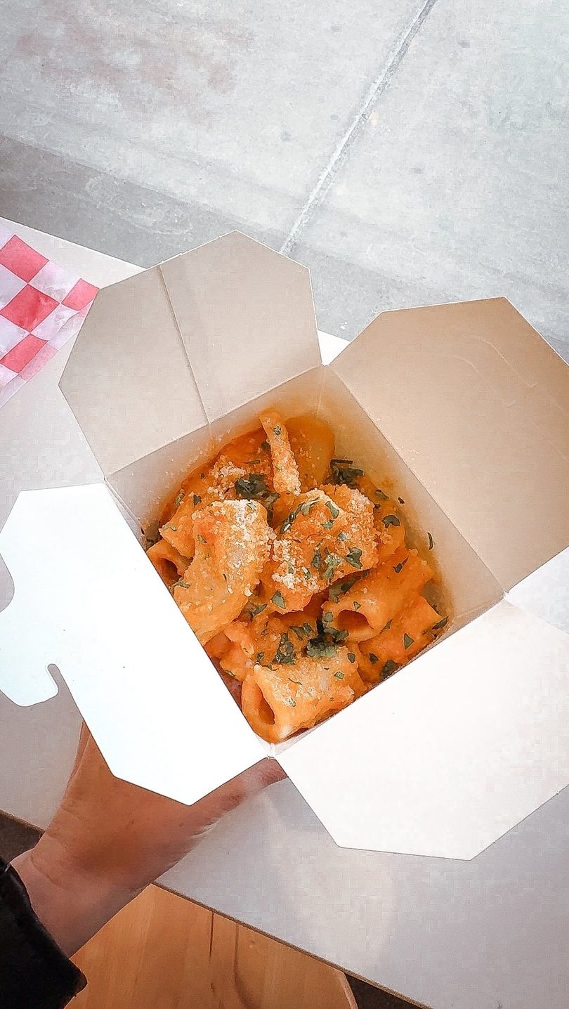 A hand holds an open white takeout box filled with rigatoni pasta in a creamy orange sauce, topped with cheese and herbs.