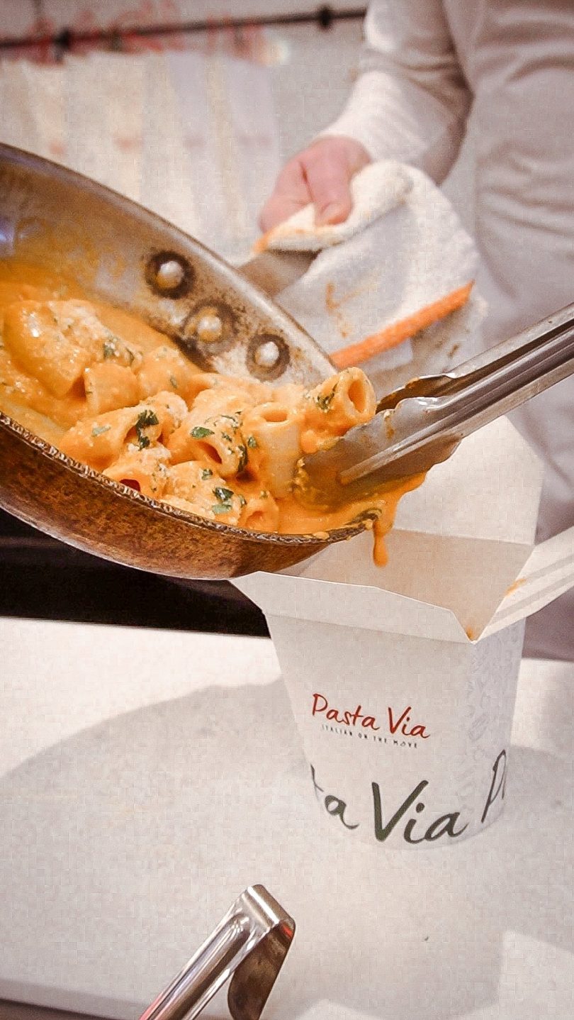 Chef using tongs to transfer creamy pasta from a pan into a "Pasta Via" takeout container.